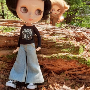 May include: A black-haired doll wearing a black long-sleeved shirt with the words "Black Sabbath" printed on it, blue jeans, and black trainers. The doll is standing in front of a large log in a woodland setting. A smaller doll with brown fur is standing behind the log.