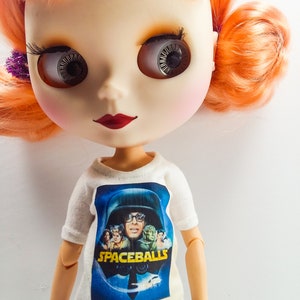 May include: A doll with orange hair wearing a white t-shirt with a Spaceballs movie poster print. The poster features a character wearing a helmet and the text "Spaceballs".