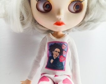 May include: A doll with white hair wearing a white long-sleeved shirt with a pink and white heart patterned pants. The shirt has a graphic of a man with the text "Close to me" and "Sweetest you".
