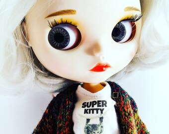 Puede incluir: Una muñeca blanca con cabello rubio que lleva una camiseta blanca con el texto "SUPER KITTY" y una chaqueta de punto multicolor.