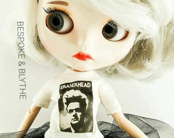 May include: A white doll with blonde hair wearing a white t-shirt with a black and white image of a man and the text "ERASERHEAD". The doll is also wearing a black tulle skirt.