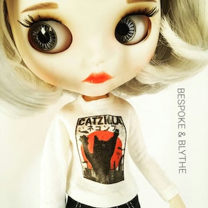 May include: A doll with large eyes, pale skin, and silver hair. The doll is dressed in a white long-sleeved top featuring a black cat graphic and the text "Catzilla" in a Japanese-style font. The doll also wears black trousers.