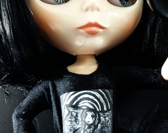 May include: A black haired doll with large eyes and a black and white graphic on a black shirt.