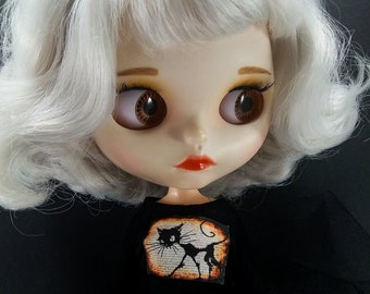 May include: A doll with white hair and a black bow, wearing a black dress with a black cat design on the front.