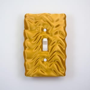 May include: A decorative light switch cover in a golden-yellow hue with a textured, quilted design. It features a white toggle switch and two screw holes. The cover's wavy pattern adds a unique touch to home decor, measured in centimetres.