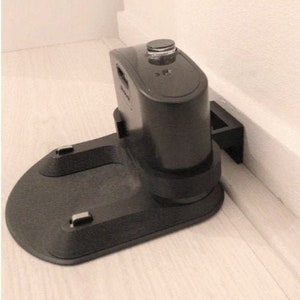 Puede incluir: Estación de carga negra para aspiradora robot. La base es de forma ovalada con dos contactos de carga empotrados. La parte vertical tiene un cuerpo cilíndrico con un pequeño botón circular en la parte superior. La unidad está montada contra una pared blanca.