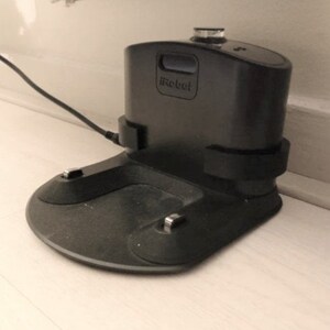 May include: A black iRobot charging station with a rounded base and a rectangular top. The iRobot logo is visible on the front. A black power cord is plugged into the side.