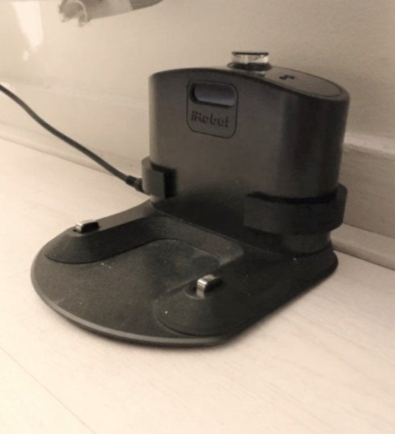 May include: A black iRobot charging station with a rounded base and a rectangular top. The iRobot logo is visible on the front. A black power cord is plugged into the side.