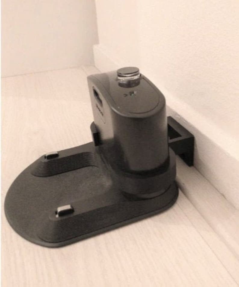 May include: A black robotic vacuum cleaner charging station. The base is oval-shaped with two recessed charging contacts. The upright portion has a cylindrical body with a small, circular button on top. The unit is mounted against a white wall.
