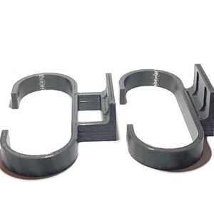 May include: Two dark gray, 3D-printed, S-shaped hooks. Each hook has a curved bottom and a flat top with a rectangular base. The hooks are designed to hang items. The hooks are made of a textured plastic material.