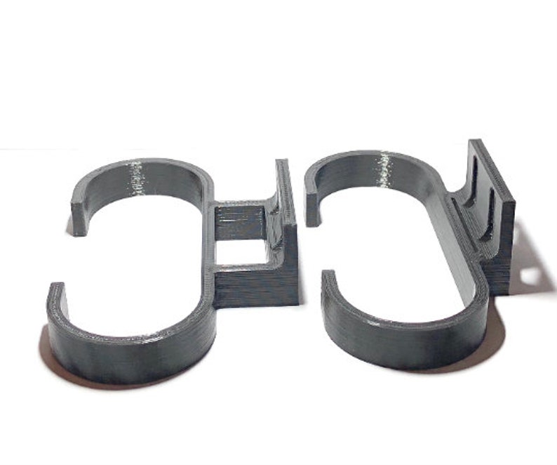 May include: Two dark gray, 3D-printed, S-shaped hooks. Each hook has a curved bottom and a flat top with a rectangular base. The hooks are designed to hang items. The hooks are made of a textured plastic material.