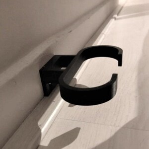 May include: A black, wall-mounted holder with a curved design. The holder is attached to a rectangular base, designed to be mounted on a wall. The curved section is open, creating a space to hold an object. The holder is made of a solid, matte material.
