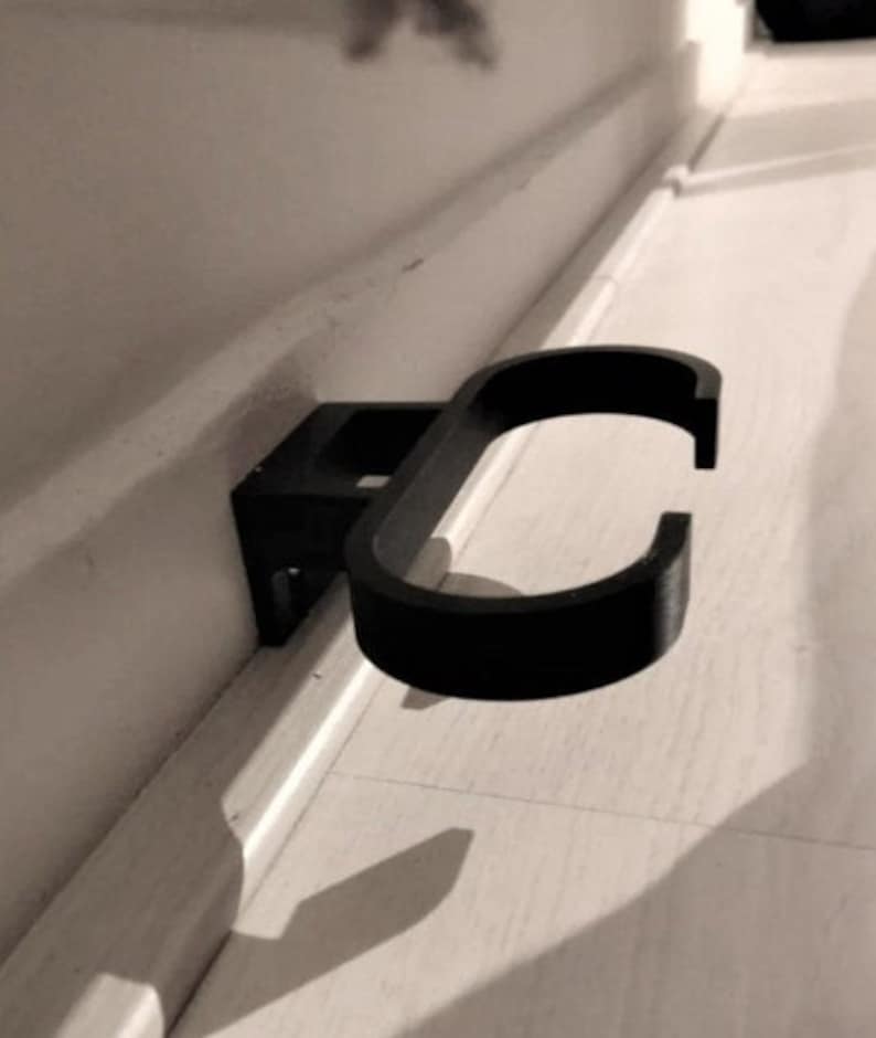 May include: A black, wall-mounted holder with a curved design. The holder is attached to a rectangular base, designed to be mounted on a wall. The curved section is open, creating a space to hold an object. The holder is made of a solid, matte material.