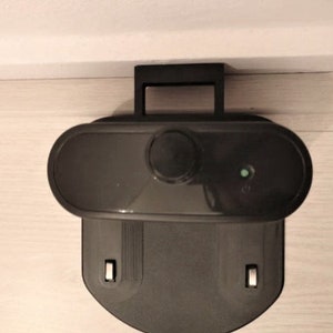 May include: A black robotic vacuum cleaner charging station. The station has a handle, an oval-shaped top with a button, and a lower section with two rectangular slots. A small green light is illuminated on the top.