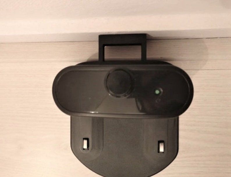 May include: A black robotic vacuum cleaner charging station. The station has a handle, an oval-shaped top with a button, and a lower section with two rectangular slots. A small green light is illuminated on the top.