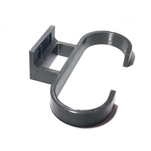 May include: A dark gray, 3D-printed wall-mounted hook. The hook features a rectangular mounting plate with two holes, a square opening, and a curved hook shape. The object is designed to hold items and is made of plastic.