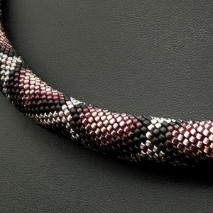 Puede incluir: Un collar de cuentas negras, plateadas y burdeos con un patrón geométrico. El collar se muestra en un maniquí negro.