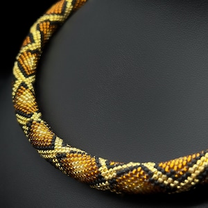 Peut inclure: Un collier de perles avec un motif de peau de serpent dans des tons d'or, de brun et de noir. Le collier est présenté sur un fond noir.