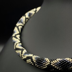 Könnte beinhalten: Eine schwarze und goldene Perlenkette mit geometrischem Muster. Die Kette besteht aus kleinen, runden Perlen, die zu einem einzigartigen Design verwoben sind.