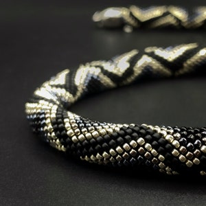May include: A black and silver beaded necklace with a chevron pattern. The necklace is made of small, round beads that are woven together in a tight, intricate pattern.