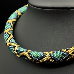 Könnte beinhalten: Eine Perlenkette mit einem geometrischen Muster in Schwarz, Gold und Grün. Die Kette besteht aus kleinen, runden Perlen, die zu einem schlangenartigen Design verwoben sind.