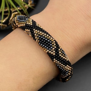 May include: A black and gold beaded bracelet. The bracelet is a cylindrical shape, made from small, tightly woven beads. The gold beads create a contrasting pattern against the black, measuring approximately 2 cm in diameter.