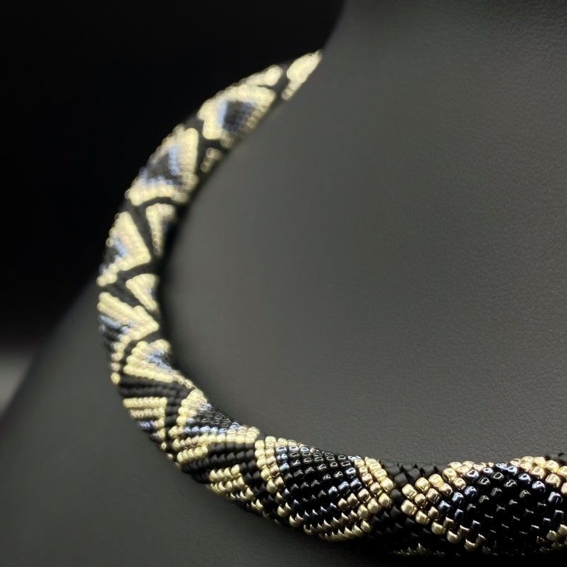 May include: A black and gold beaded necklace with a geometric pattern. The necklace is made of small, round beads that are woven together to create a unique design.
