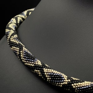 May include: A black, gold, and silver beaded necklace with a geometric pattern. The necklace is made of small beads and is twisted into a spiral shape.