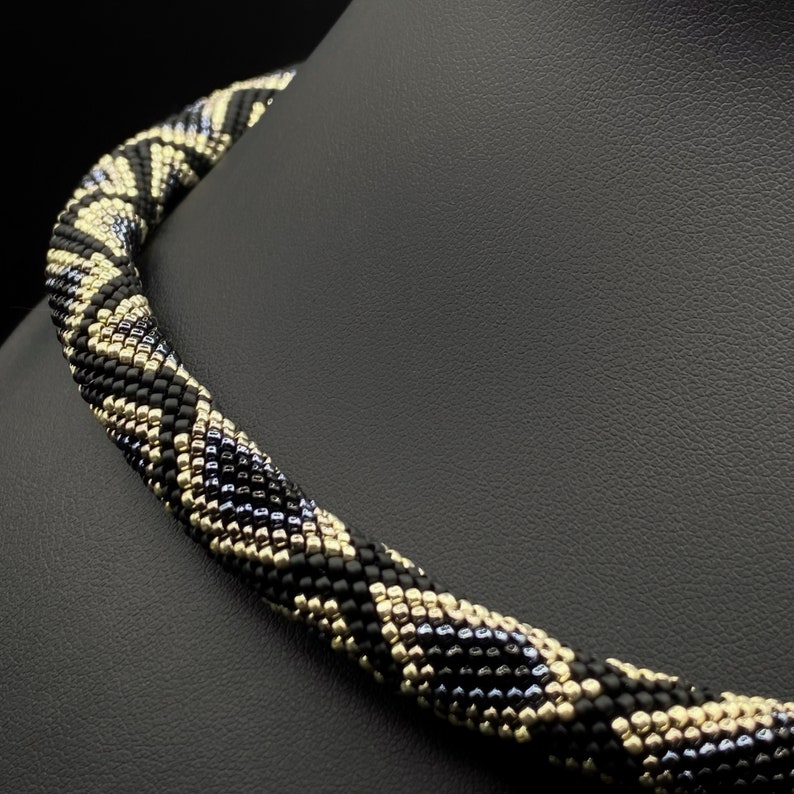 May include: A black, gold, and silver beaded necklace with a geometric pattern. The necklace is made of small beads and is twisted into a spiral shape.