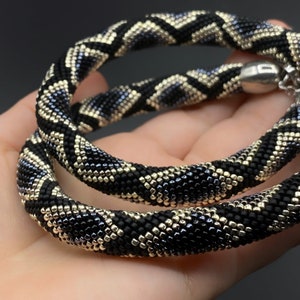 May include: A black, gold, and silver beaded necklace with a geometric pattern. The necklace is made of small, square beads and is approximately 18 inches long.