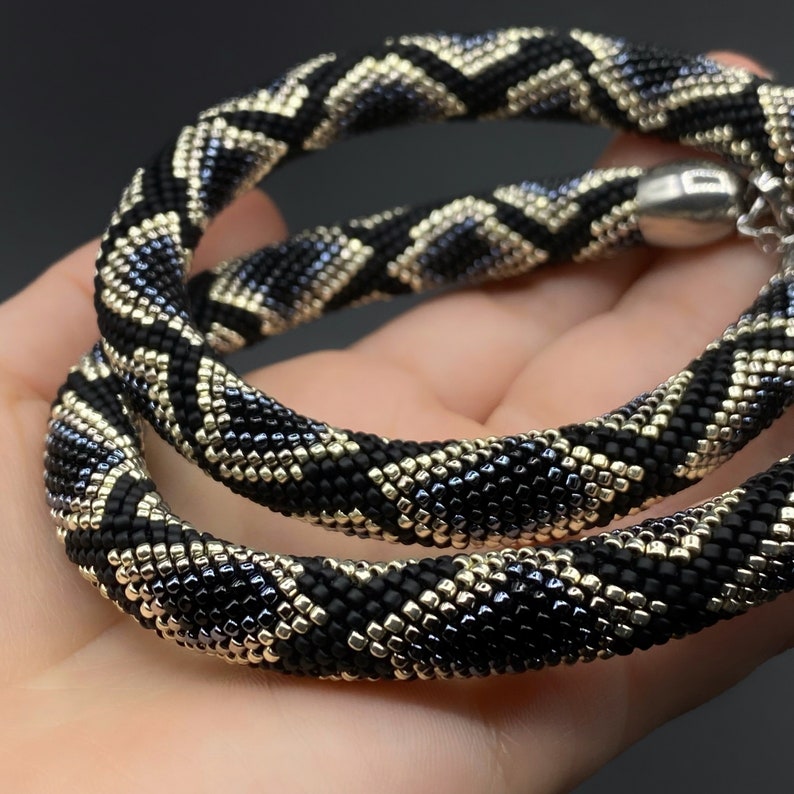 May include: A black, gold, and silver beaded necklace with a geometric pattern. The necklace is made of small, square beads and is approximately 18 inches long.