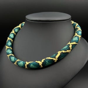 May include: A beaded necklace in shades of teal and gold, displayed on a black mannequin bust. The necklace features a repeating pattern of teal beads with gold accents, creating a textured, eye-catching design. The necklace is a statement piece.
