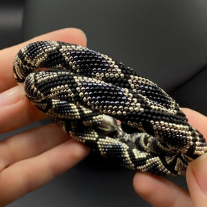 May include: A black and gold beaded necklace with a geometric pattern. The necklace is made of small, round beads that are woven together to create a unique and stylish design.