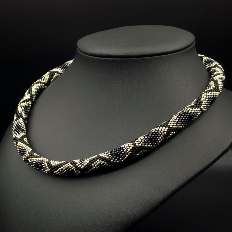 May include: A black and white beaded necklace with a geometric pattern. The necklace is on a black display stand.