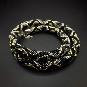 May include: A black and gold beaded necklace with a geometric pattern. The necklace is made of small, square beads and has a silver clasp.