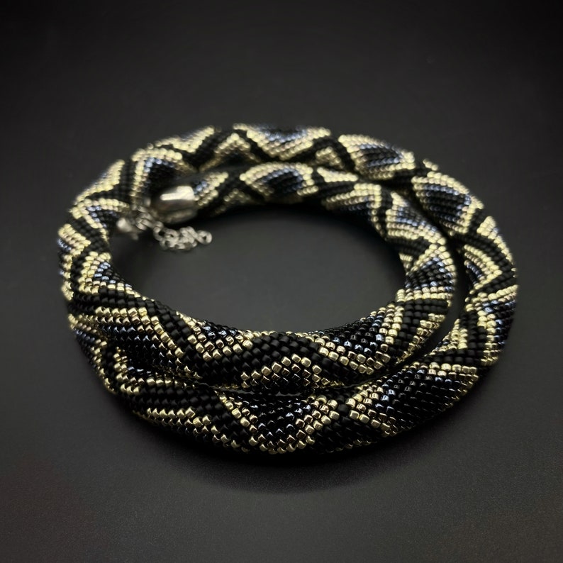 May include: A black and gold beaded necklace with a geometric pattern. The necklace is made of small, square beads and has a silver clasp.