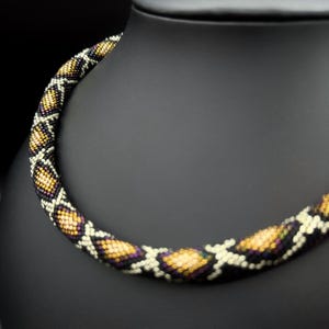 Op de afbeelding: Een kralenketting met een slangachtig patroon. De ketting heeft een herhalend ontwerp van gele, paarse en witte kralen tegen een zwarte achtergrond. De ketting wordt gepresenteerd op een zwarte mannequin.