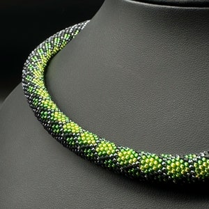 Op de afbeelding: Een zwarte en groene kralenketting met een geometrisch patroon. De ketting is gemaakt van kleine, ronde kralen die in een herhalend patroon van zwart en groen zijn gerangschikt.