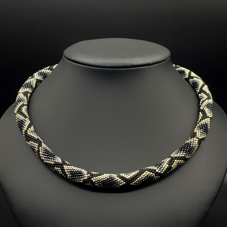 May include: A black and white beaded necklace with a geometric pattern. The necklace is on a black mannequin.
