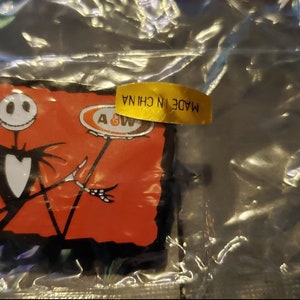 May include: An A&W root beer themed Jack Skellington pin. The pin features Jack Skellington from the Nightmare Before Christmas movie holding a root beer mug. The pin is red, black, and white. The pin is packaged in a clear plastic bag with a yellow label that reads "Made in China."