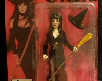 May include: Elvira Witch Variant action figure in a plastic package. The figure is wearing a black dress and holding a broom. A smaller black witch hat is also included. The packaging has the Elvira logo and the Figures Toy Co. logo.