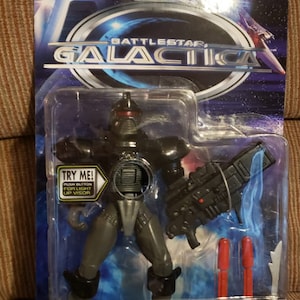 May include: Battlestar Galactica Cylon Centurion action figure in its original packaging. The figure is grey and black with a red visor, and includes a missile blaster and two red missiles. The packaging features the Battlestar Galactica logo.