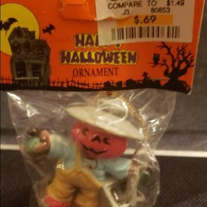 May include: A Halloween ornament featuring a pumpkin-headed scarecrow wearing a white hat, light blue shirt, and yellow pants. The ornament is packaged in a clear plastic bag with an orange background that reads "Happy Halloween Ornament."