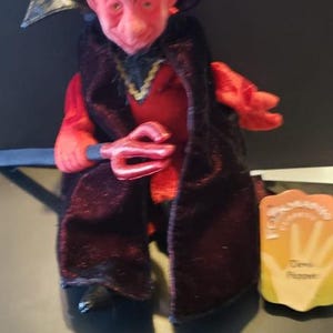 May include: A red devil puppet with a black cape and a trident. The puppet has small horns and a mischievous expression. A small tag is visible on the right side of the puppet.