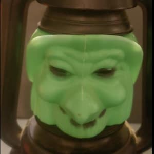 以下が含まれることがあります： 緑色の光る顔をした黒いプラスチック製のハロウィーンランタン。顔は大きく歯の見える笑顔をしていて、大きな黒い目があります。