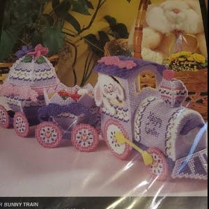 May include: A Bucilla plastic canvas craft kit for a bunny train featuring a purple and pink locomotive and two attached carriages. The train is decorated with floral patterns and the text Happy Easter. The kit includes 10 mesh plastic canvas.