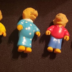 May include: Four small figurines of bear characters. One wears yellow overalls over a yellow shirt, another wears a blue polka-dot nightgown, and the other two wear red shirts and blue pants. All have yellow feet and hands.