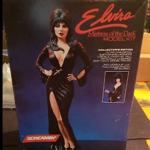 May include: A model kit box featuring Elvira, Mistress of the Dark. The box has a dark blue background with Elvira in a black dress. Text on the box includes "Collector's Edition" and "Over 18" tall". The box is from Screamin'.