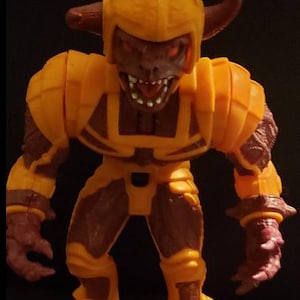 May include: A detailed action figure of a monster with a yellow helmet and armor. The figure has a brown face with red eyes and sharp teeth. The monster has horns and is posed in a menacing stance.