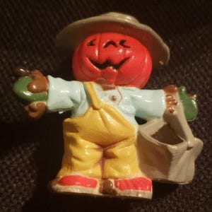 May include: A ceramic figurine of a smiling pumpkin-headed scarecrow wearing a light blue shirt, yellow overalls, and a gray hat. The scarecrow is holding a green apple and a gray bag. Red shoes complete the outfit.
